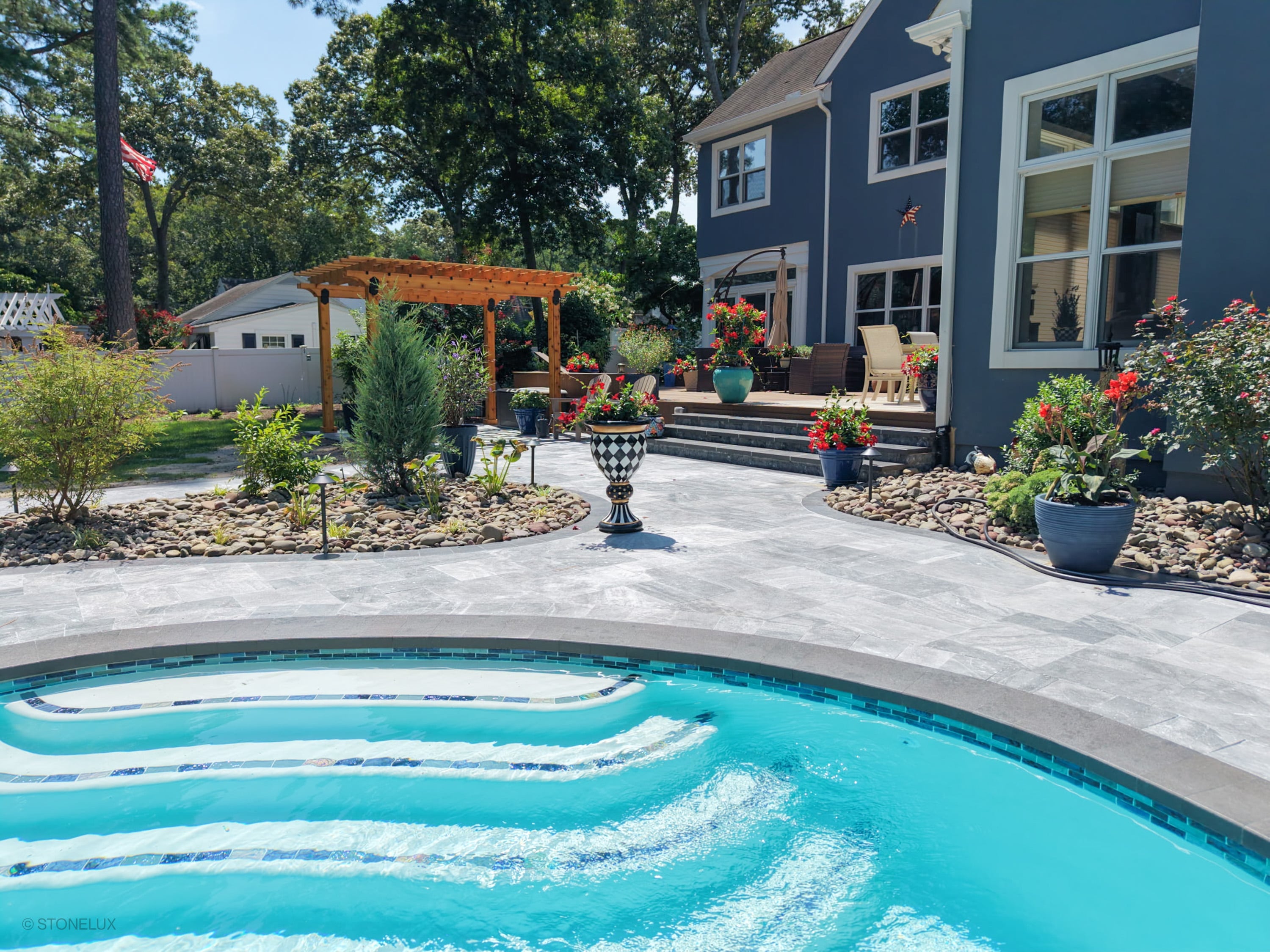 Aegean Grey Marble Pool Retreat in Salisbury, MD, showcases elegant black basalt coping around a vibrant blue pool. The modern design features natural stone pavers, lush landscaping with potted plants, a wooden pergola, and cozy outdoor seating, creati...