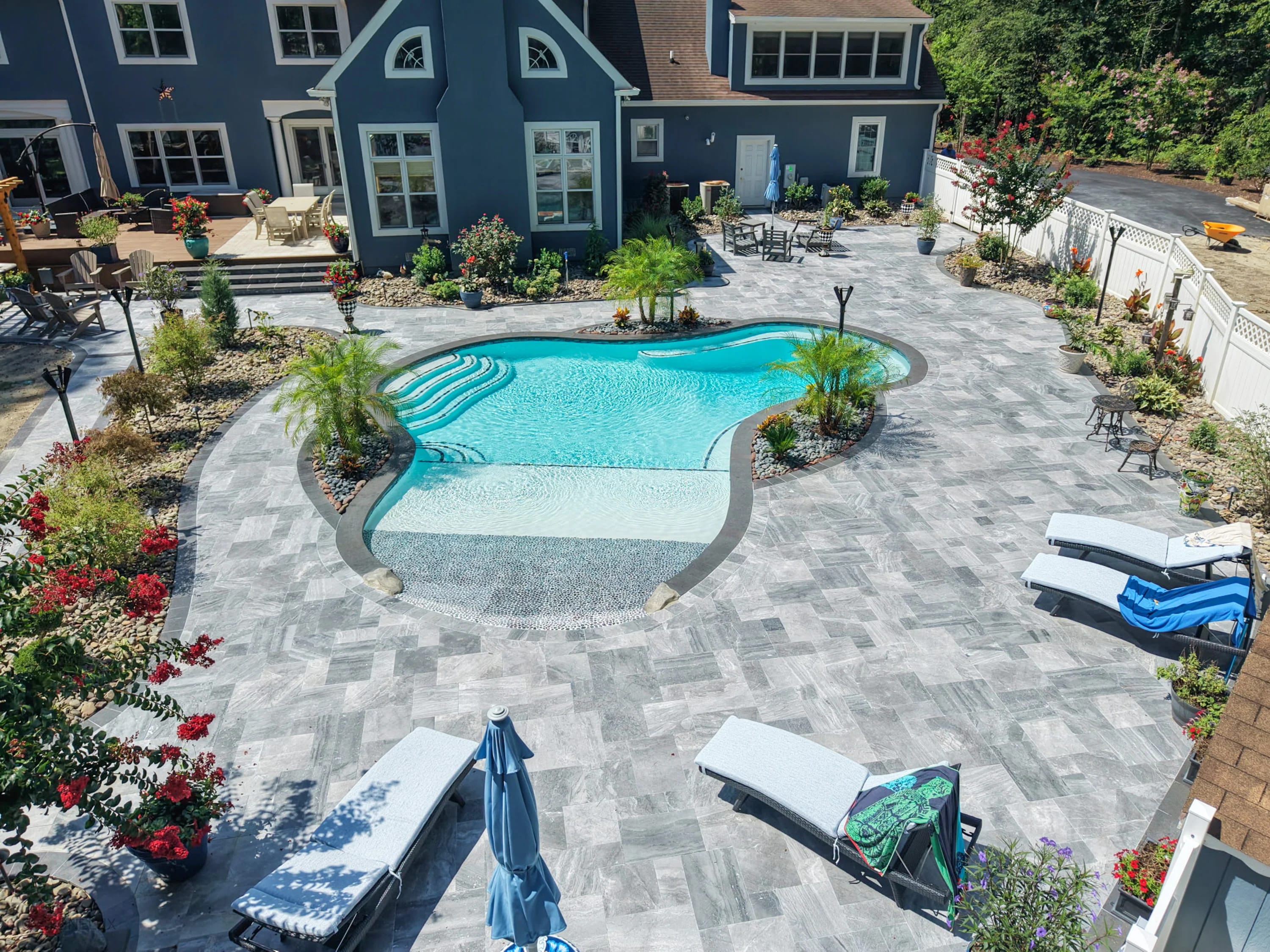 Aegean Grey Marble Pool Retreat in Salisbury, MD, features sleek grey marble pavers and black basalt coping. The modern design surrounds a sparkling blue pool, complemented by lush landscaping, red blooms, and white lounge chairs, creating a serene bac...