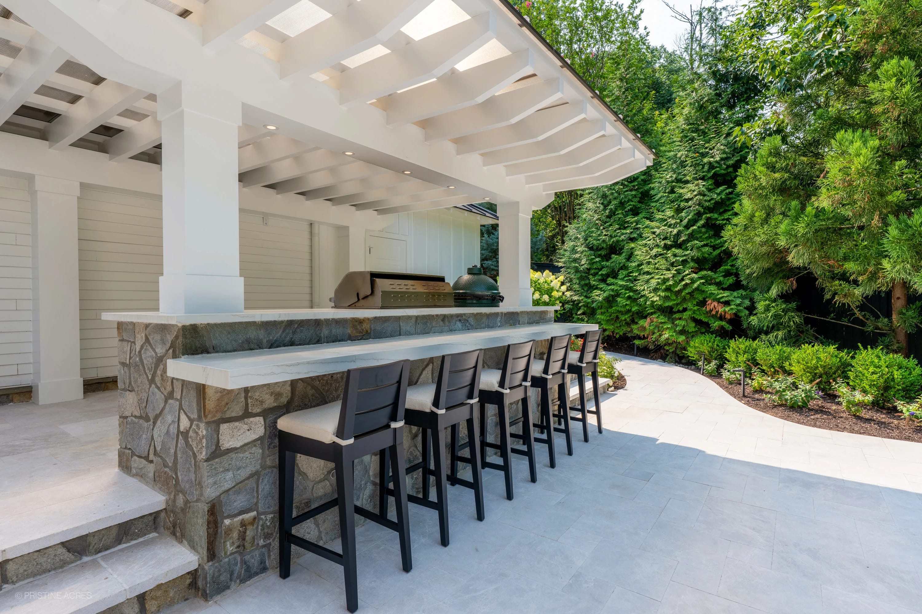 Elegant outdoor living space in McLean, VA, featuring Pera Cream Marble from STONELUX. Modern design with a stone bar, white pergola, and sleek black chairs. Lush greenery surrounds, enhancing the serene patio atmosphere.