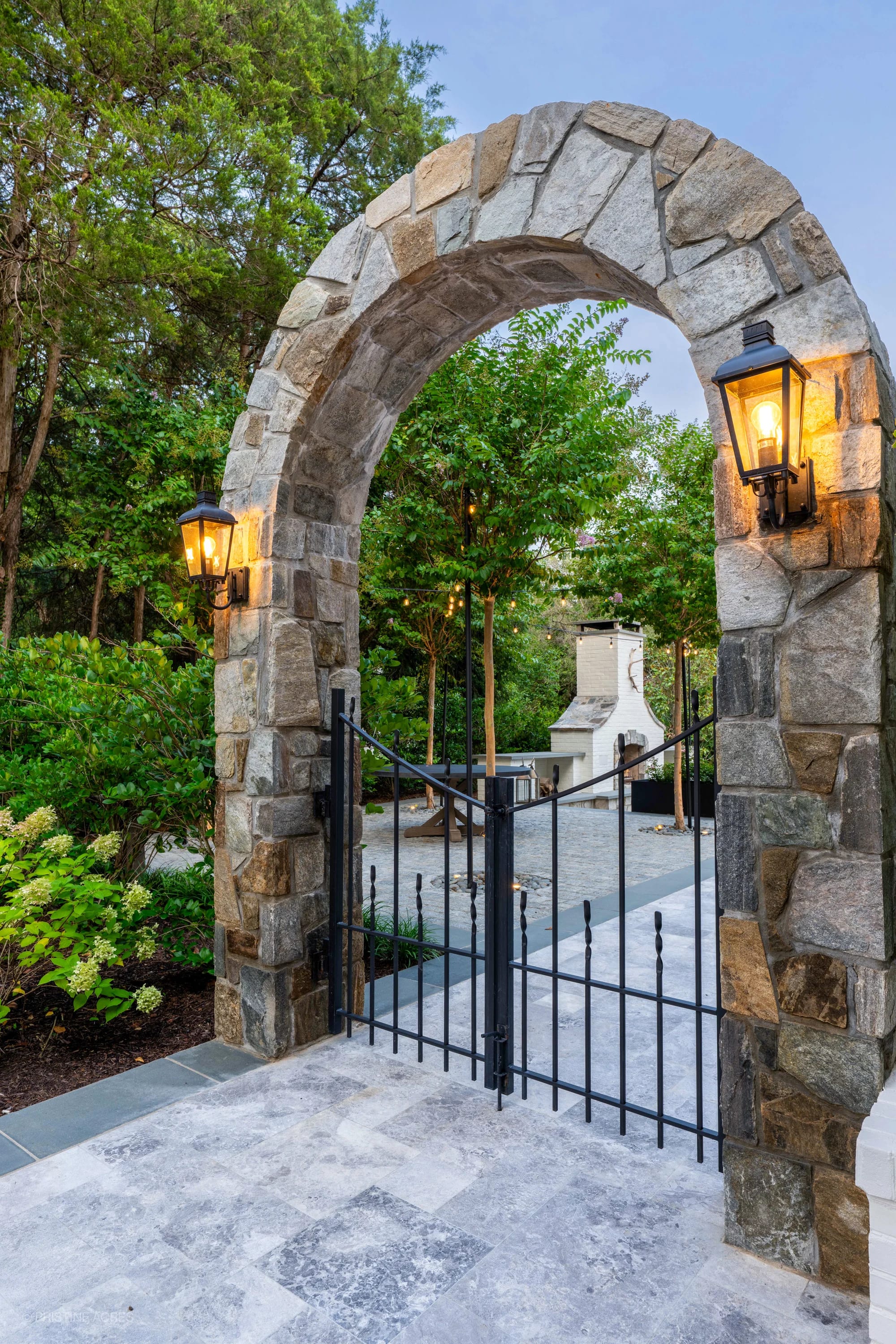 Elegant hardscape project in McLean, VA featuring Tundra Grey Marble pavers. Stone archway with lanterns leads to a lush garden patio. Bluestone coping complements natural greenery and a modern outdoor fireplace, creating a serene, upscale ambiance.