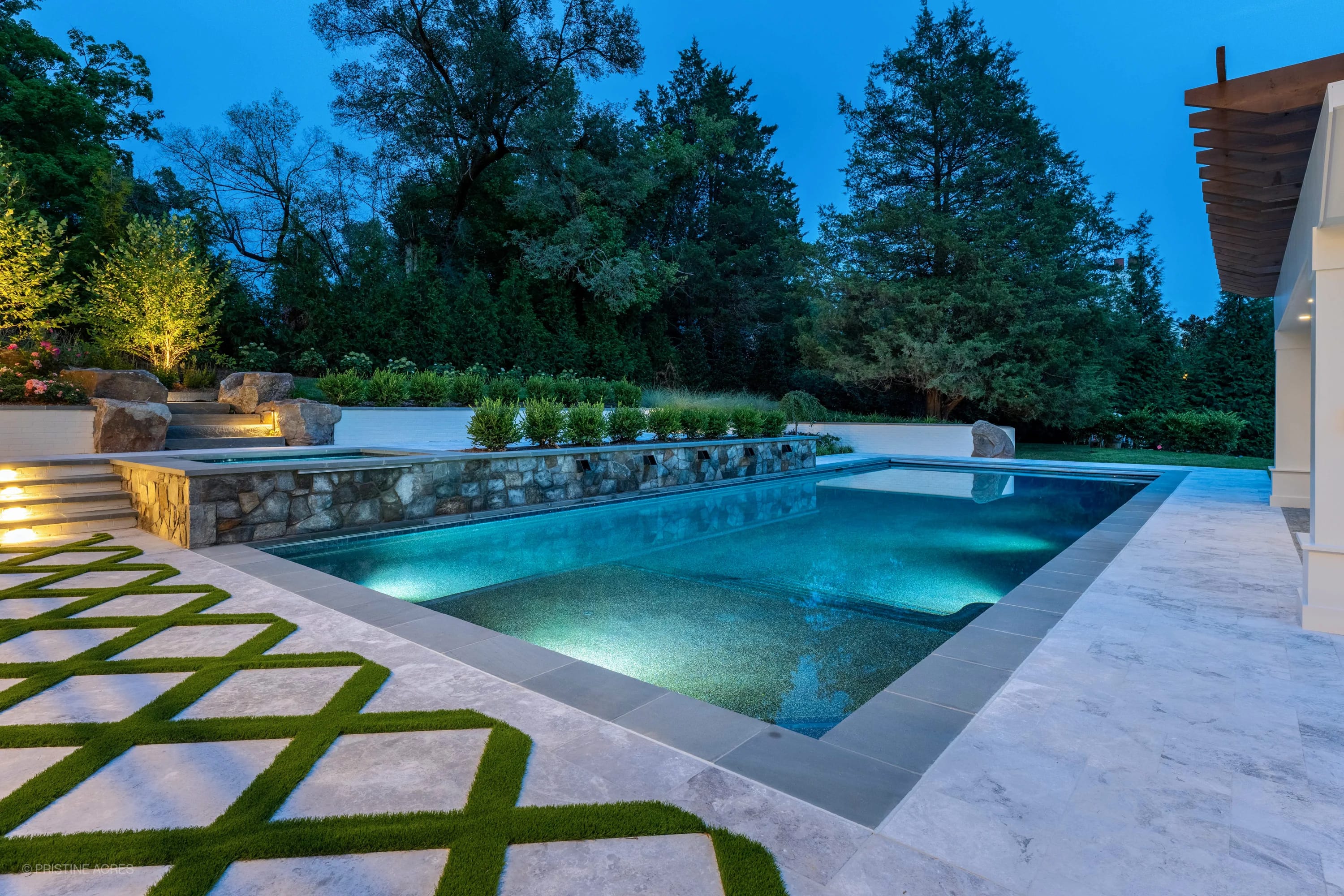 Luxurious pool deck in McLean, VA featuring Tundra Grey Marble pavers and Bluestone coping. The modern design includes a turquoise pool, stone accents, lush landscaping, and elegant outdoor lighting, creating a serene backyard oasis.