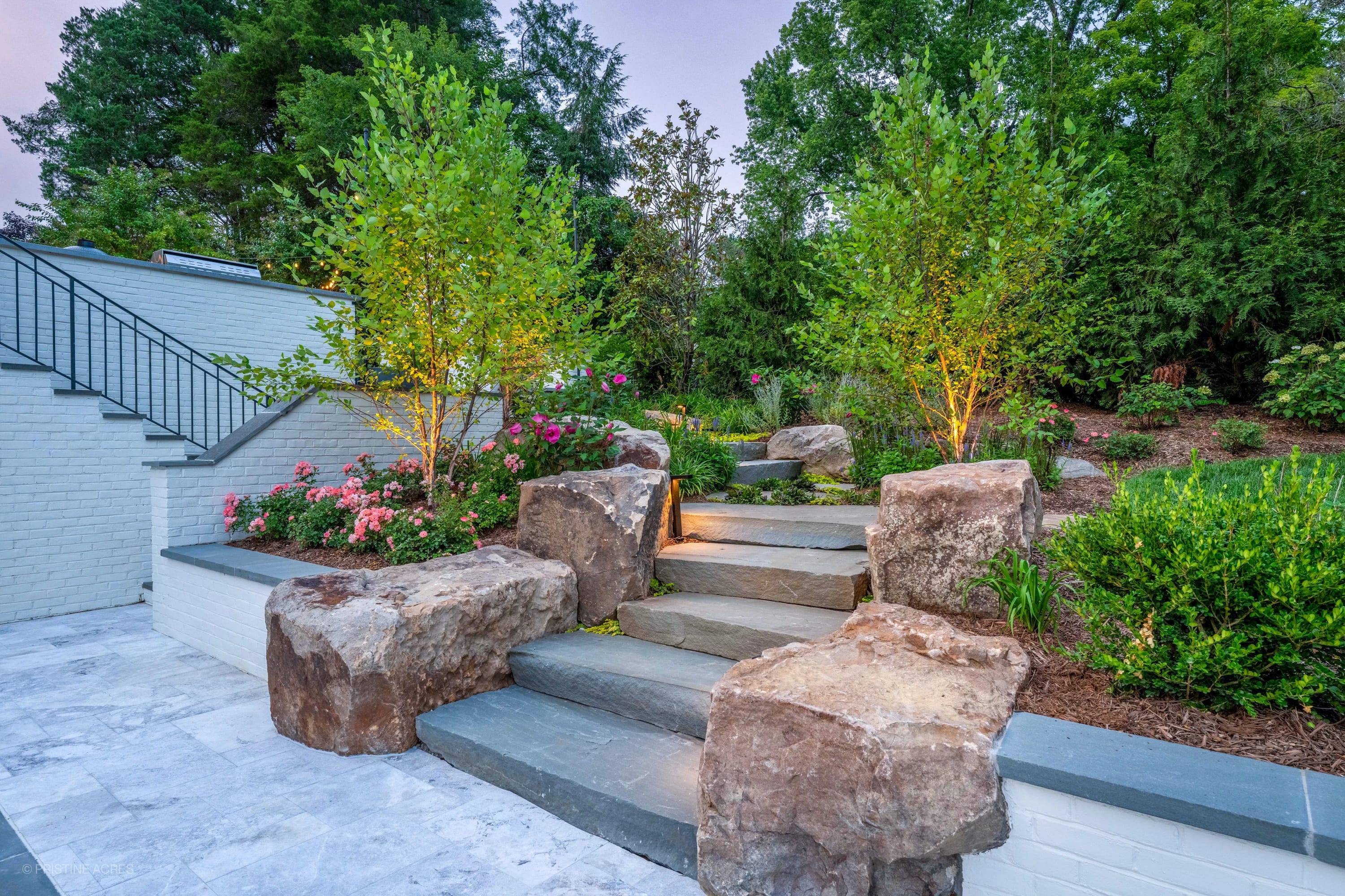 Tundra Grey Marble pool deck in McLean, VA, featuring Bluestone coping. The modern design includes lush landscaping with vibrant flowers and greenery. The elegant stone steps and ambient lighting create a serene backyard atmosphere. Perfect for luxury ...