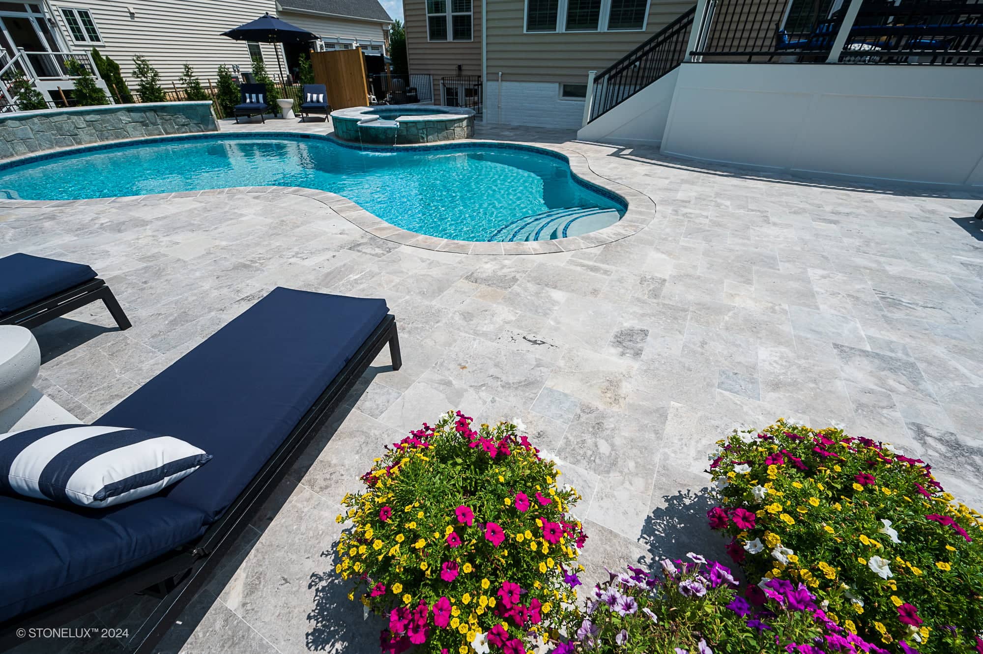 A backyard with a swimming pool and hot tub, surrounded by elegant Tundra Grey Marble tiles. Colorful flowers brighten the scene, while lounge chairs with dark cushions sit poolside. Houses and a shaded seating area complete this Project in Ashburn VA.