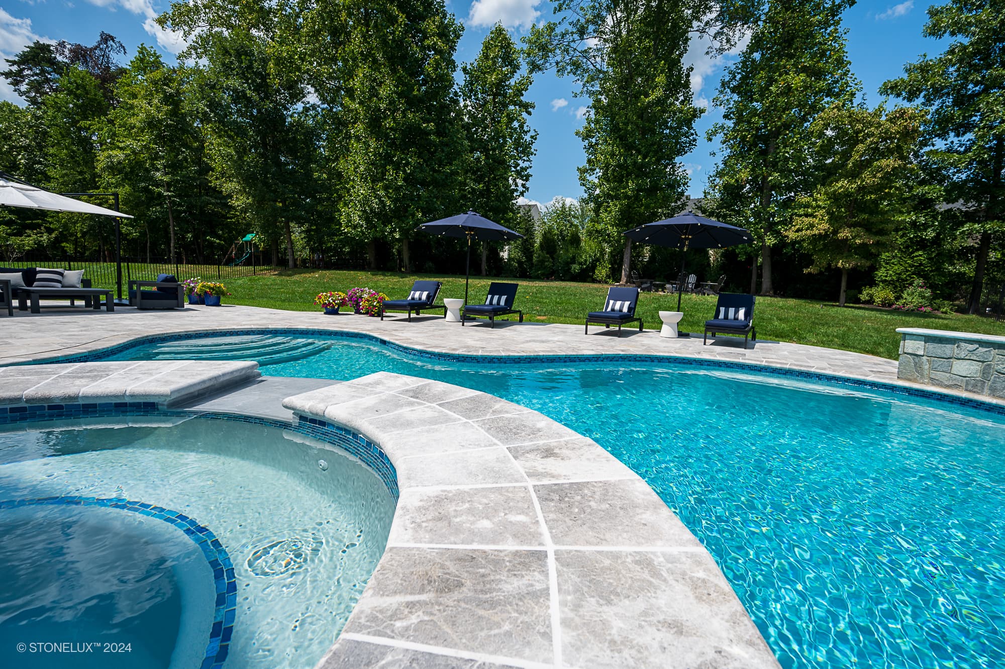 A luxurious backyard pool and built-in hot tub are surrounded by Tundra Grey Marble stone decking, lounge chairs, umbrellas, and lush green trees—perfect for a Pool and Patio Project in Ashburn VA under a sunny, blue sky.