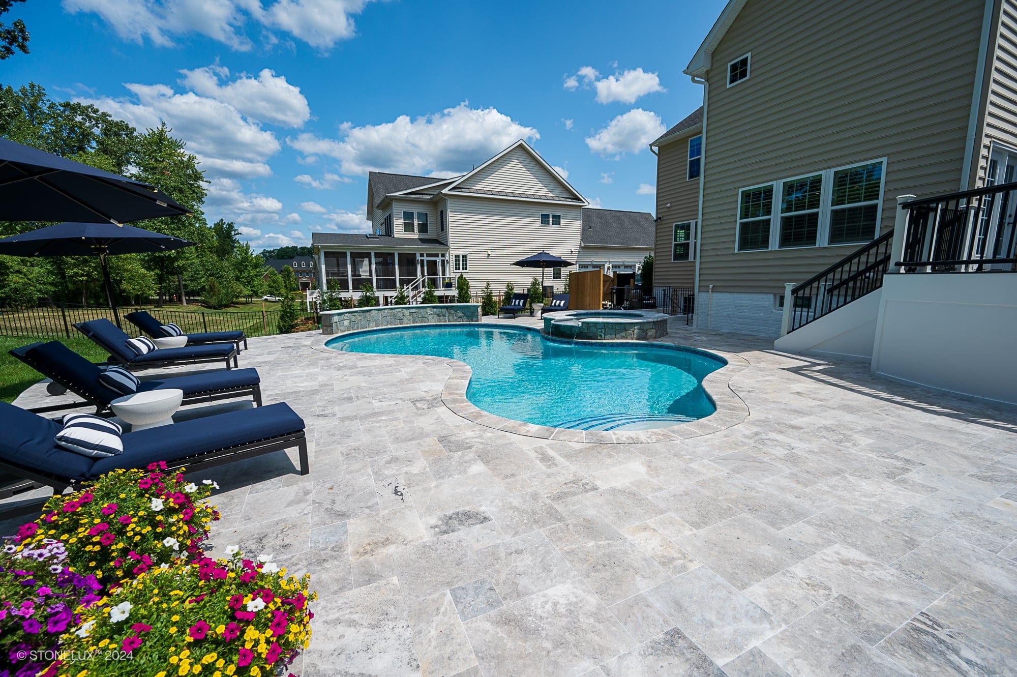 A backyard in Ashburn, VA features a curvy swimming pool and patio project with Tundra Grey Marble, lounge chairs with striped pillows, large umbrellas, blooming flowers, modern houses, and a blue sky with scattered clouds.