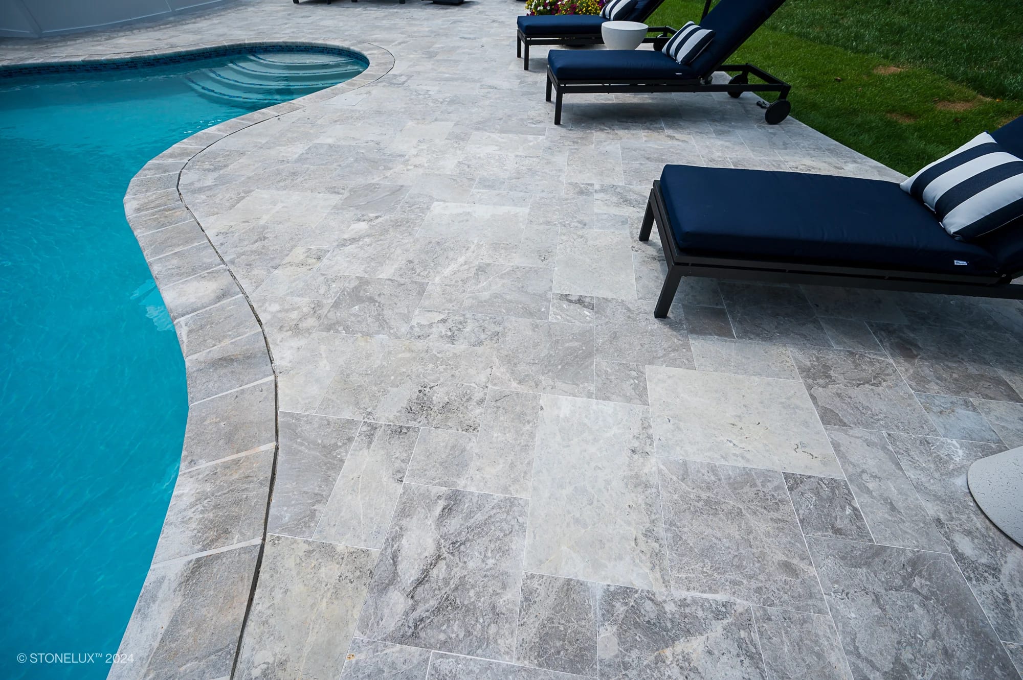 Curved-edge swimming pool with Tundra Grey Marble tile deck, two navy blue lounge chairs with striped pillows, and a patch of green grass in the background. Pool and Patio Project in Ashburn VA.